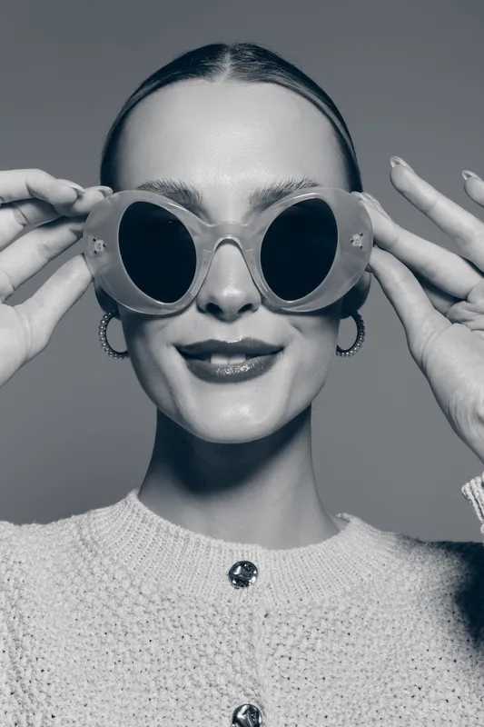 Young Caucasian woman with fair skin wearing stylish large round sunglasses and hoop earrings smiles confidently in studio. Concept of fashion, beauty, and accessories portrait.