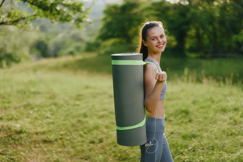 Young woman carrying yoga mat in outdoor setting, expressing joy and vitality, showcasing healthy lifestyle in nature