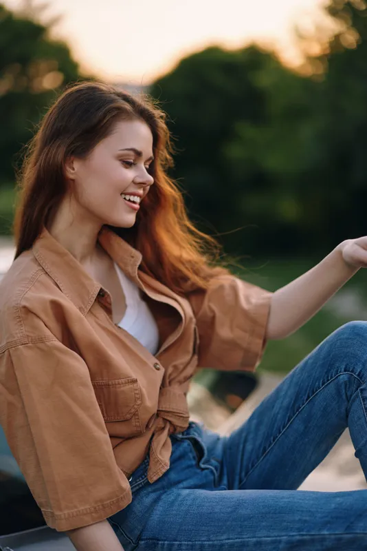 Young woman with long hair wearing a stylish beige shirt and jeans, enjoying the sunset outdoors, radiating happiness and warmth in a natural setting.