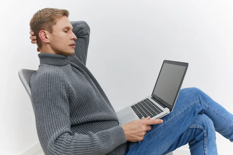 Relaxed young man with blond hair wearing a cozy gray turtleneck sweater, sitting back with eyes closed and resting, holding a laptop in a bright minimalistic room, casual style, comfort concept.