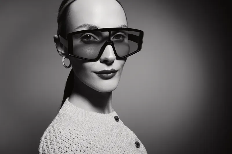 Portrait of a stylish white woman with pale skin wearing large black sunglasses and a white knitted sweater on grey background.