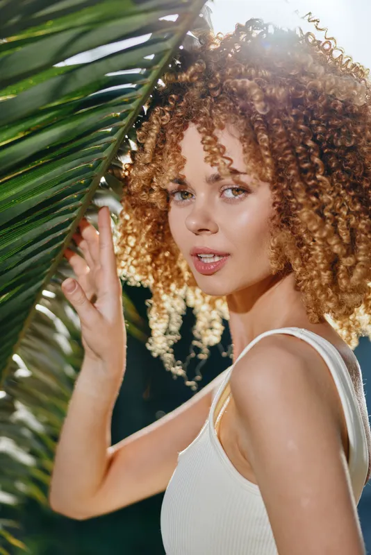 Curly woman with natural hair wearing white tank top stands behind green palm leaves, basking in warm sunlight with soft expression and smooth glowing skin.