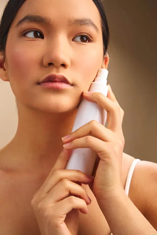 asian woman holds a skincare bottle near her face, exuding calm confidence as she pursues a gentle beauty routine in soft, natural light.