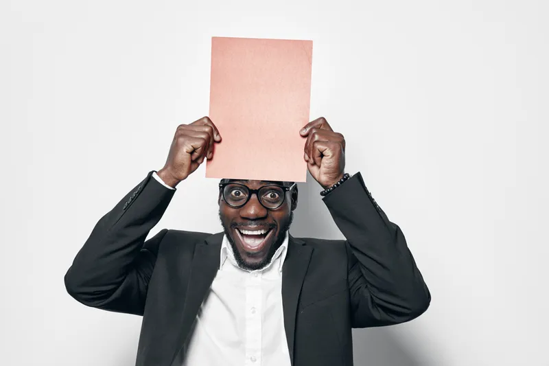 African man with dark skin and glasses holding blank paper above head smiling in studio. Black male in suit and white shirt with joyful expression on white background for business or education.