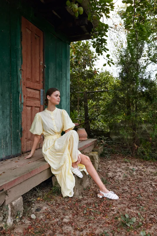 Woman sits on wooden steps outside a green house wearing a pale yellow dress and white sandals in a sunlit garden setting
