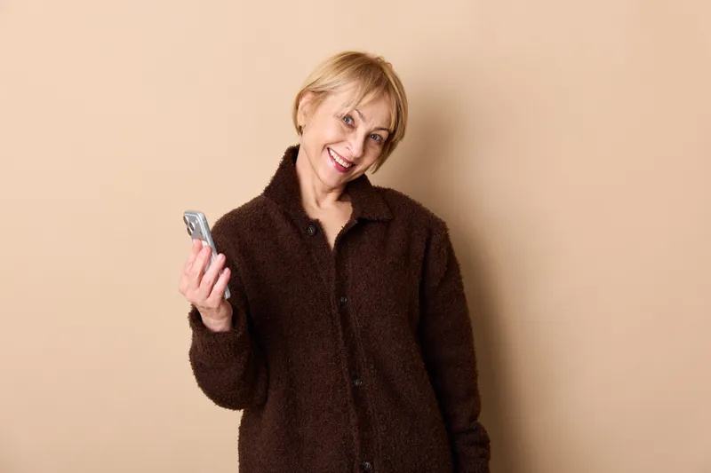 Smiling woman with smartphone, holding phone in hand, wearing brown winter coat, indoor portrait on beige background, relaxed communication mood, copy space