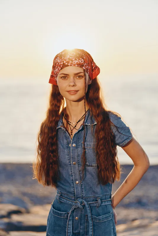 young woman wearing red bandana and denim dress standing by the sea at sunset with long hair and warm sunlight on her face