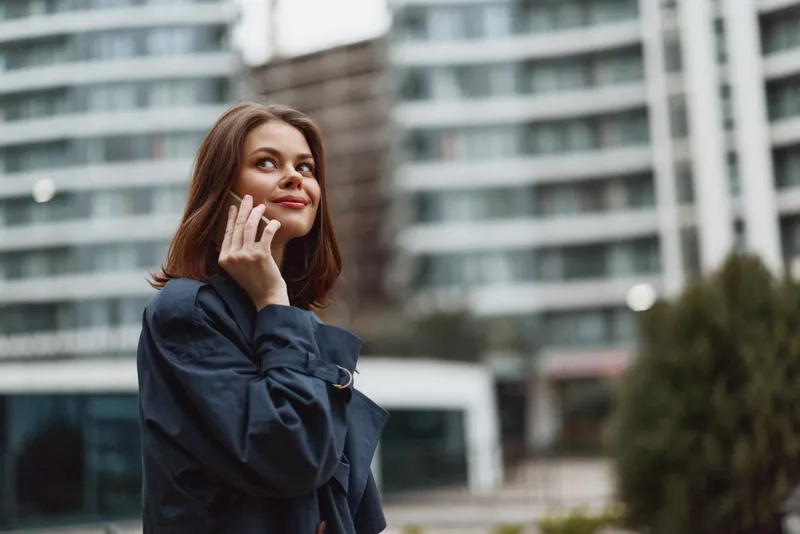 Young woman with long brown hair talks on a smartphone outdoors near modern buildings with a thoughtful expression on her face.