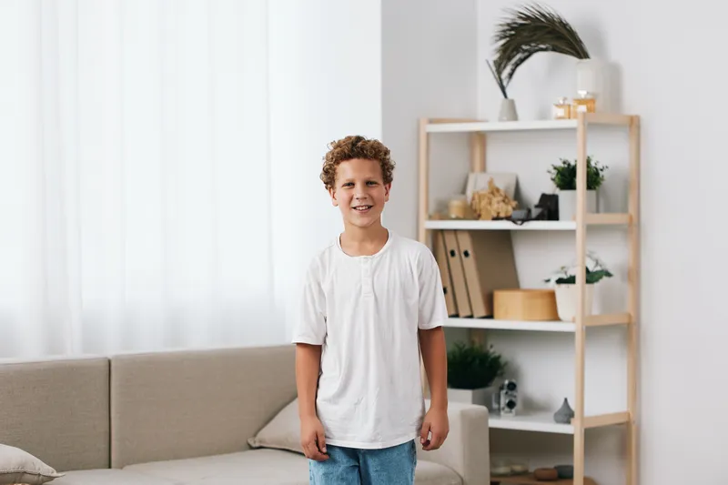 Smiling boy stands indoors in a bright, minimalist living room with white shirt and jeans, creating a candid portrait with warm natural light, soft cushions, and greenery in the background.