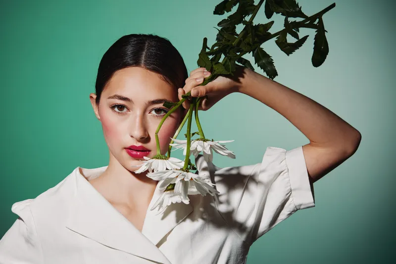 Young woman with smooth skin and dark hair holds white daisies near face on green background wearing white blouse. Minimalist beauty creative fashion concept with soft natural light and shadow.