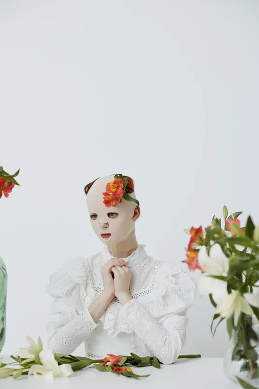 Serene woman with a floral mask in a minimalistic setting, showcasing emotions of tranquility and beauty, surrounded by fresh flowers and soft colors