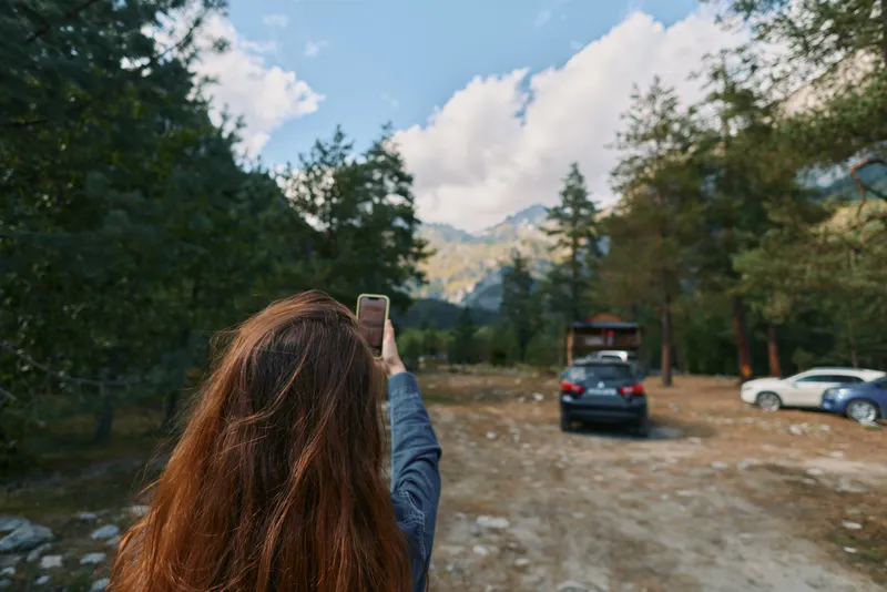 A person with long hair stands in a forest clearing, holding a smartphone to photograph a distant car on a dirt road, as tall trees frame the sunny outdoor scene.