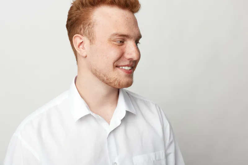 Smiling young man with red hair and beard wearing white shirt looks sideways. Studio portrait on light background. Concept of happiness, casual style, and positive emotions.