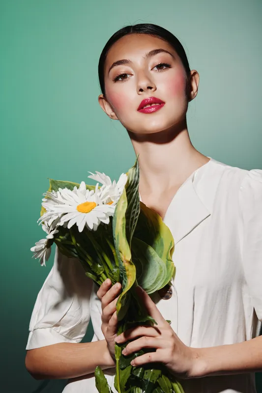 Young woman with glossy lips holds bouquet of white daisies. Her flawless skin and minimal white top highlight fresh beauty concept. Soft studio lighting on muted background.
