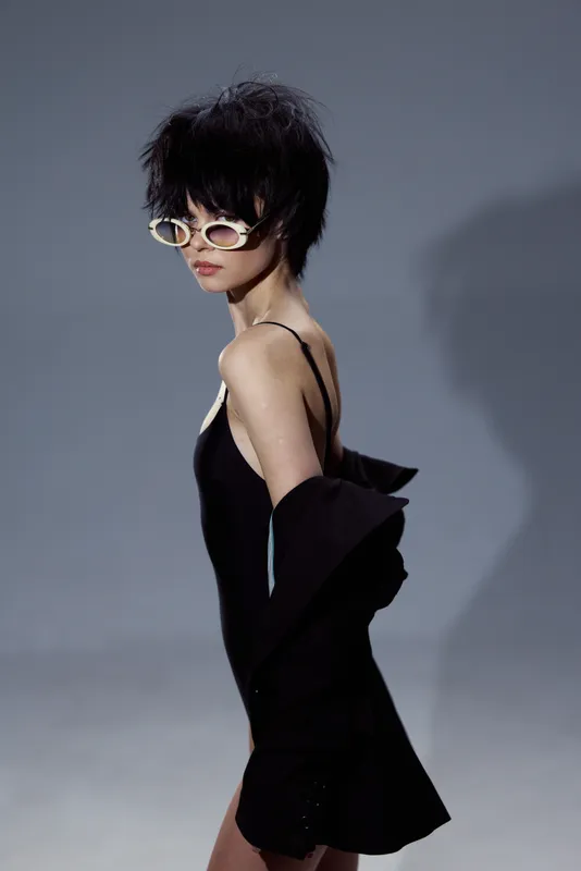 Stylish woman in black dress and sunglasses posing against gray background with confident expression and modern hairstyle