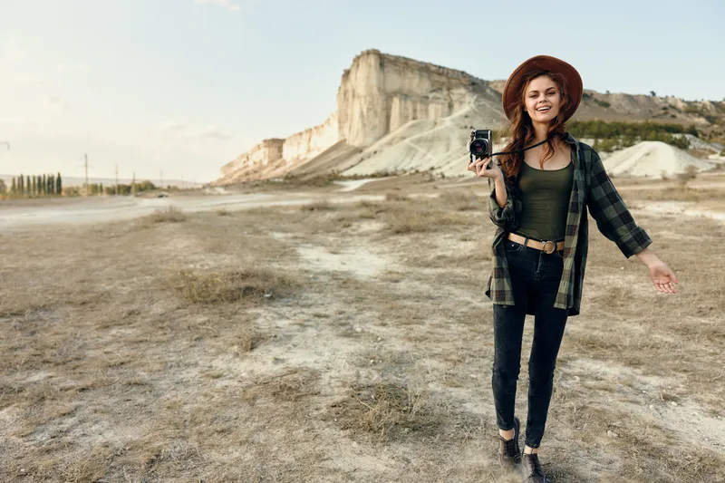 Женщина-фотограф в клетчатой рубашке и шляпе снимает природу на камеру, находясь на открытом поле