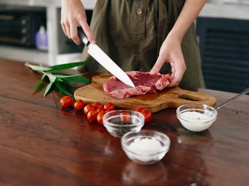 Женщина с ножом в руке режет сырое мясо для жарки на кухне