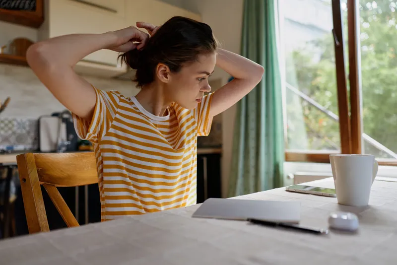 One woman sits at a home desk in a striped shirt, fixing her hair as natural light streams through a window, creating a calm, focused moment for a lifestyle shot.