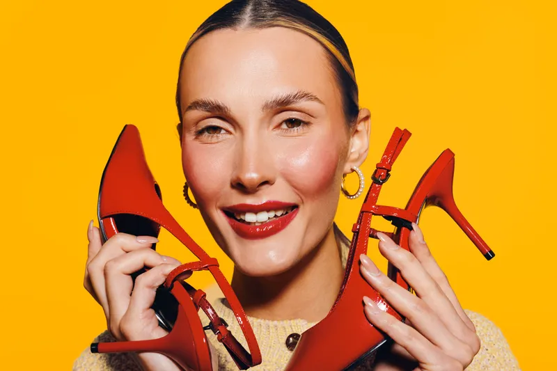 Smiling woman with red lipstick holds red high heel shoes near face on bright yellow background. Concept of fashion, style, beauty, and vibrant color contrast in studio lighting.
