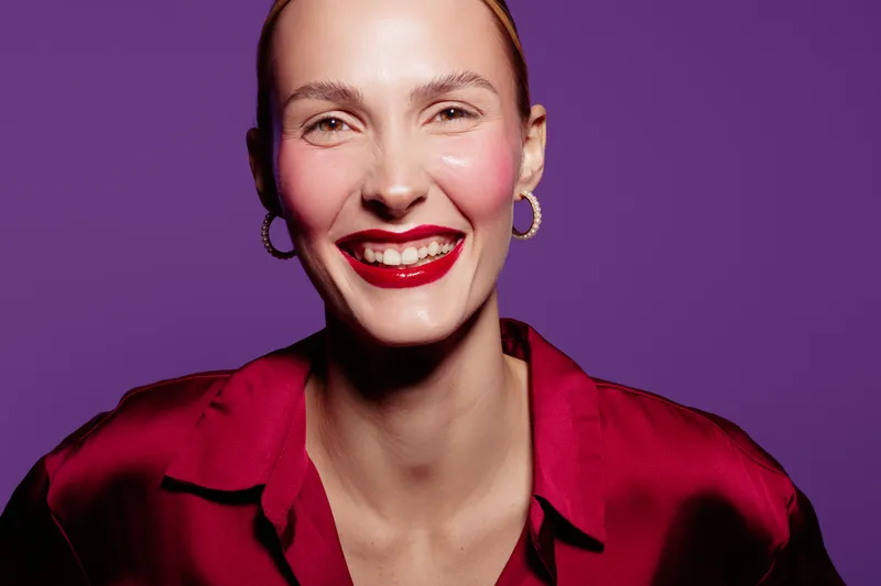 Smiling woman with red lipstick and hoop earrings wears a red satin blouse against purple background. Close-up portrait with bright makeup and joyful expression. Concept of beauty and fashion.