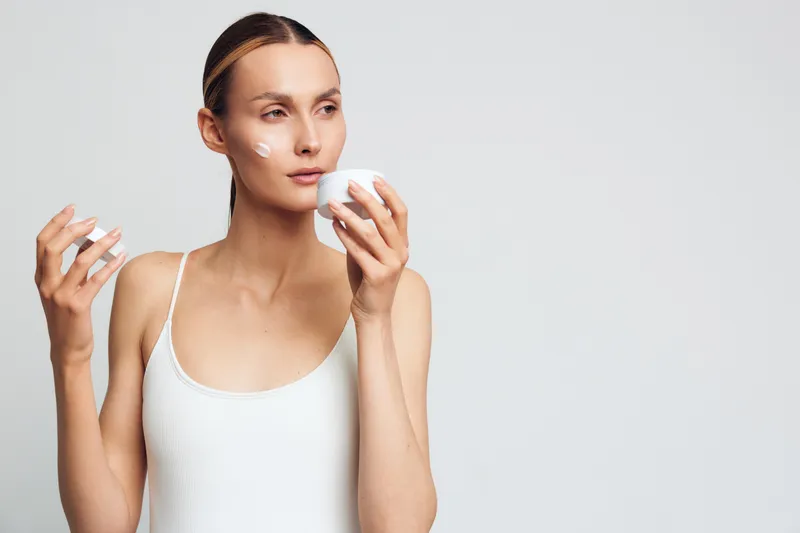 Young woman in white tank top applies face cream with cotton pad looks away with calm expression isolated on light background skincare beauty routine concept.