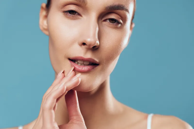 Closeup portrait of young woman touching face with fingers, smooth skin, natural makeup, soft lighting, blue background, calm expression, skincare concept, beauty routine.