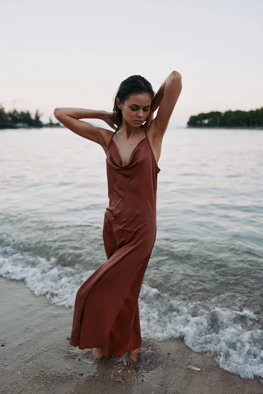 A woman in a rust brown dress stands at the shoreline, waves lap at her feet, a relaxed pose, wind in hair, with soft sunset light highlighting the silhouette.