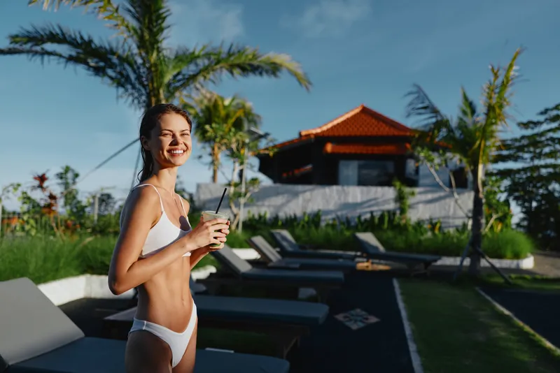 Young woman in a white bikini smiles poolside at a tropical resort, holding a glass and enjoying a sunlit moment as palm trees and a resort terrace frame the scene, she looks relaxed and confident by