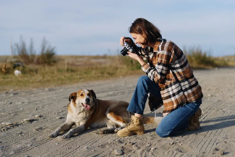 Photographer kneels beside a dog outdoors, capturing a candid moment on a sunny dirt road, plaid shirt and boots, wide landscape and quiet connection with nature