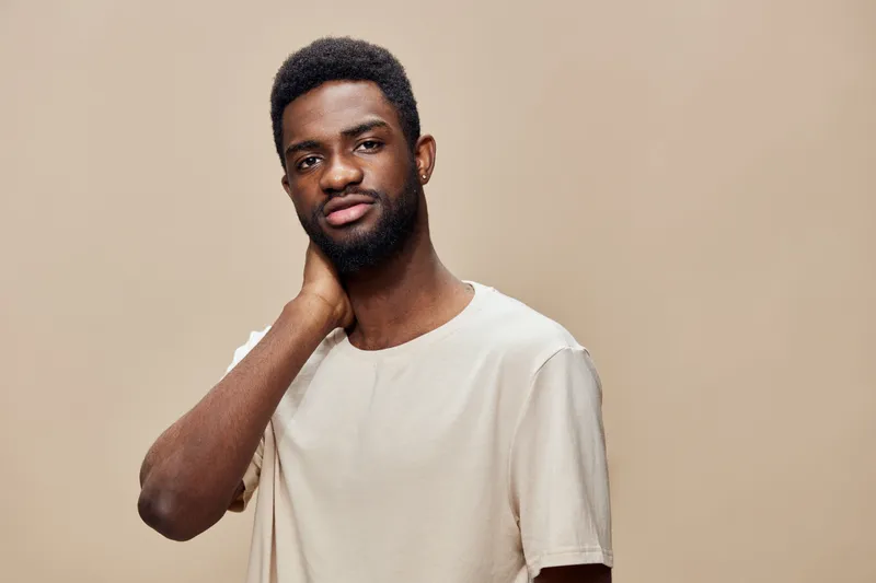 Confident young man with dark skin, wearing a casual beige t shirt, showcasing a thoughtful expression against a warm neutral background