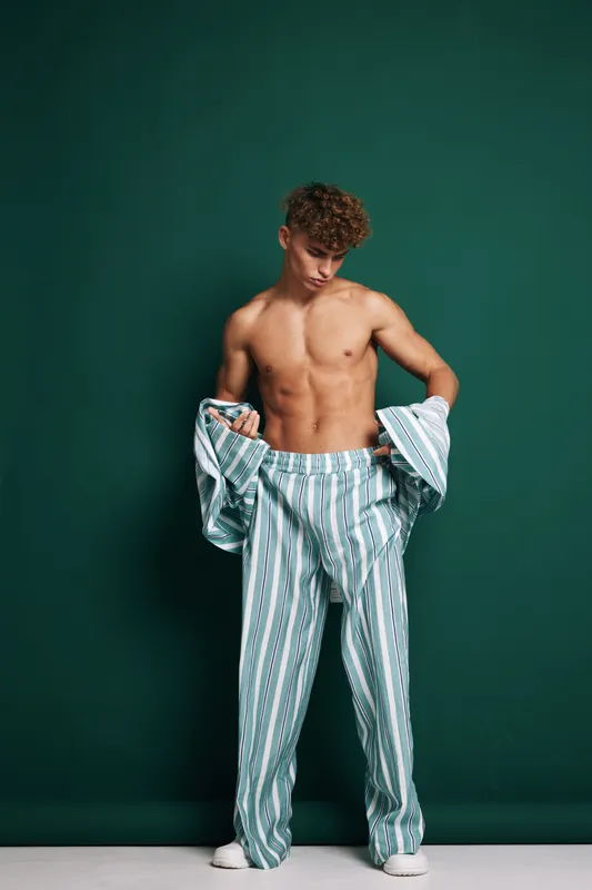 Young man with curly hair wearing striped loose pants and holding matching shirt, posing shirtless against green background in studio for fashion or lifestyle concept.