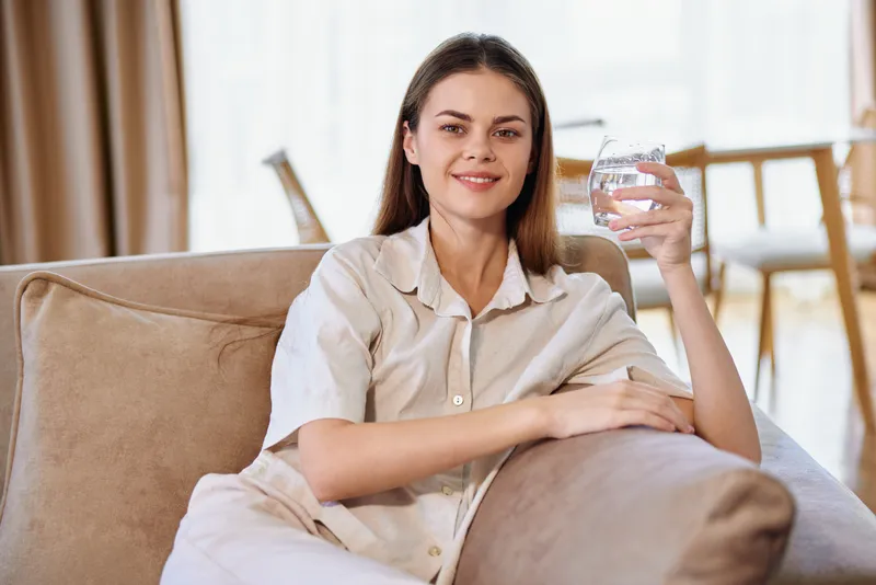 Woman on sofa smiling and holding a glass of water at home, a relaxed portrait of a young adult enjoying comfort and wellbeing on a cozy couch in a bright living room.