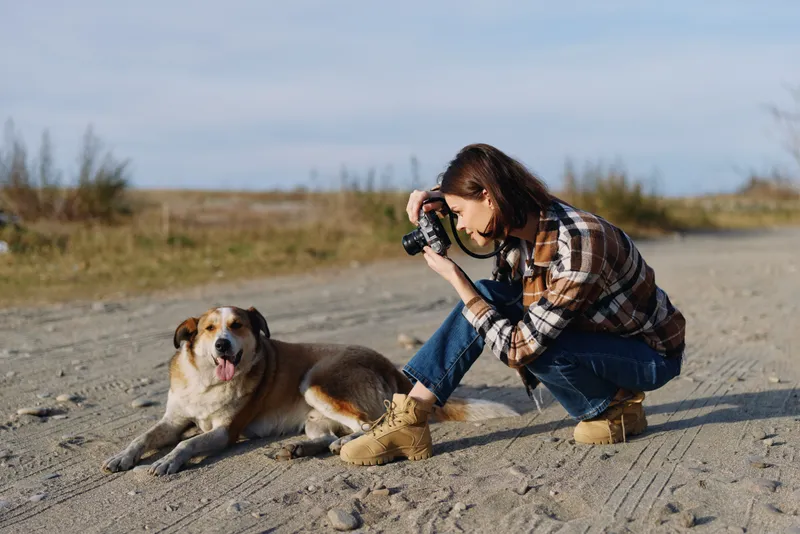 A photographer kneels on a dirt path to frame a dog with a camera, capturing a candid outdoor moment, natural light, and simple joy in a rustic landscape.