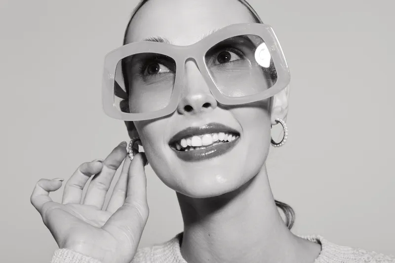 Young white woman with pale skin wearing oversized glasses and hoop earrings smiles while adjusting earring. Studio portrait with bright soft lighting.