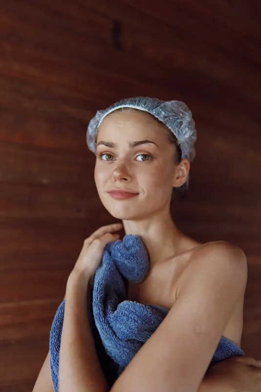 young woman with shower cap wrapped in blue towel standing in wooden sauna, calm facial expression, natural light, warm toned cozy atmosphere