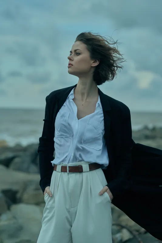 woman, beach, windswept, fashion, blazer, white shirt, trousers, belt, outdoor, sea, horizon, rocky shore, wind, hair, confident pose, coastal scenery