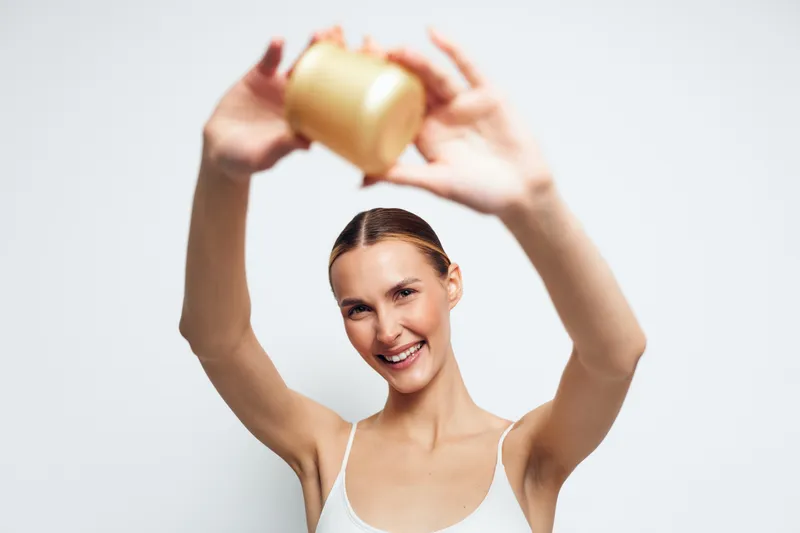 Woman beauty skincare serum bottle smiling studio white background wellness self care, holding golden container above head, cheerful expression, soft studio light, minimal aesthetic