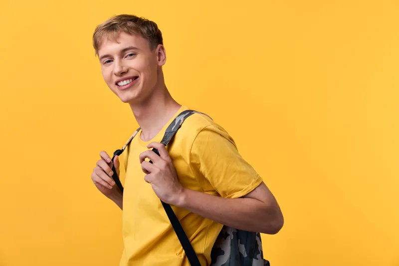 Smiling young man with backpack against a bright yellow backdrop, casual style, ready for study or travel, relaxed pose and upbeat mood, everyday wardrobe and cheerful energy.