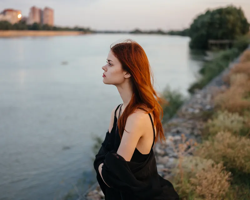 Рыжеволосая женщина в черном платье стоит у реки на природе