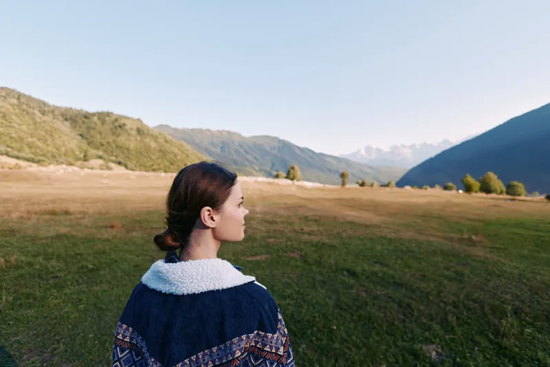 Woman stands in meadow looking at mountain landscape, portrait capturing nature and distant peaks in a serene outdoor scene, peaceful valley view for travel and reflection.
