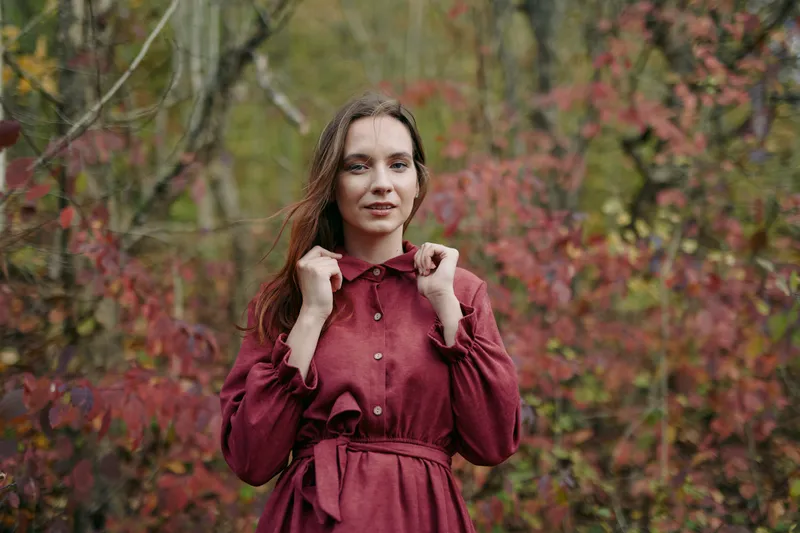 A candid portrait features a young woman in a burgundy dress amid autumn foliage, emphasizing authenticity and credibility with natural expression and relaxed posture, suitable for lifestyle and