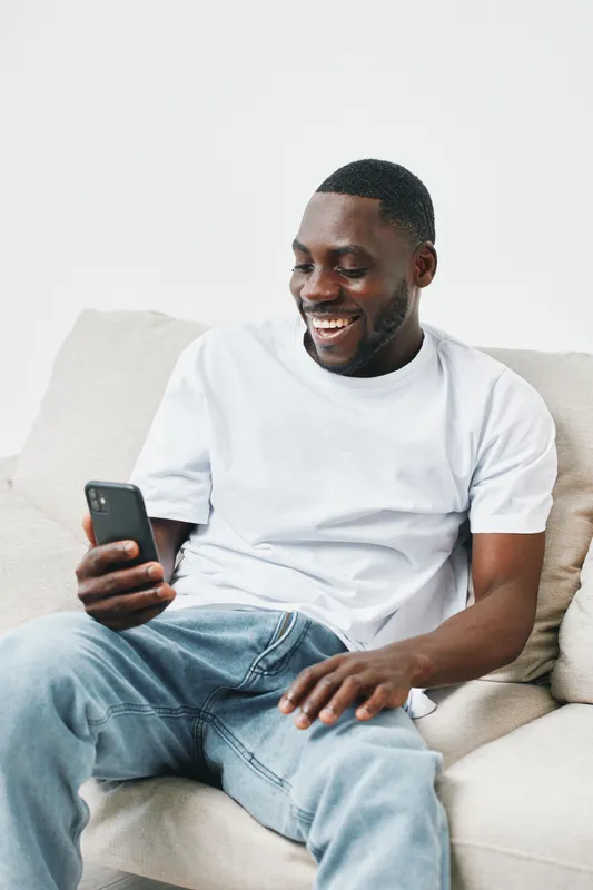 Smiling young Black man enjoying his time on a smartphone while sitting on a cozy couch, showcasing relaxed vibes and casual fashion