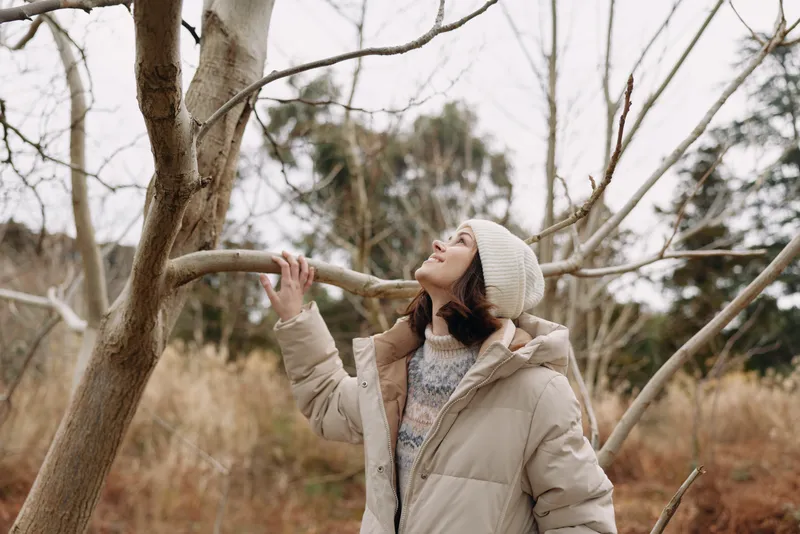 Woman in a beige winter coat and beanie reaches a branch in a bare tree landscape, capturing calm outdoor moments and a quiet seasonal mood during a cool day.