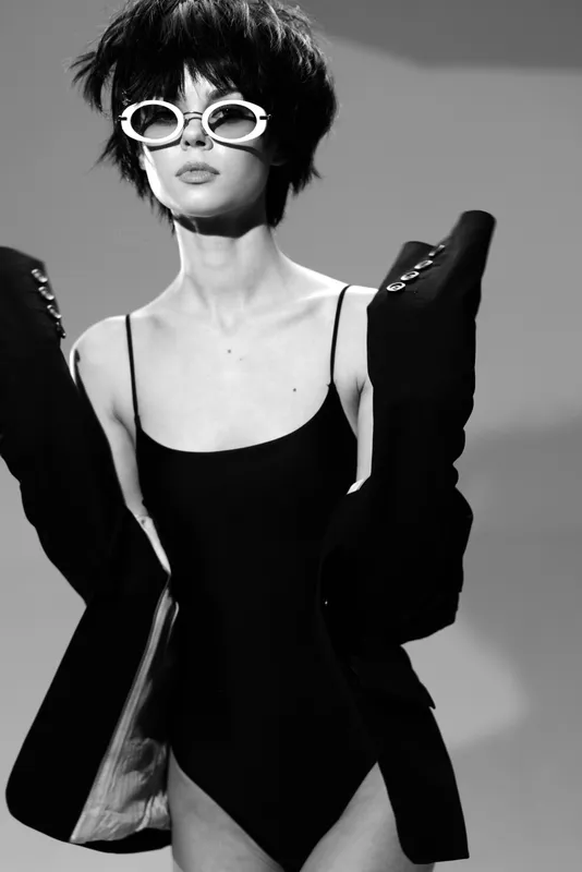 Stylish woman in black swimsuit and sunglasses posing confidently in black and white.