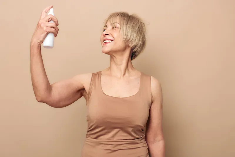 Mature woman in beige tank top sprays mist on face, eyes closed and smiling. Skin care routine, hydration, wellness, and self-care concept with soft neutral background and natural light.