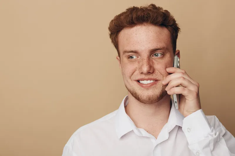 Smiling young man with red hair and freckles talks on smartphone. Casual white shirt and beige background create a warm, friendly atmosphere. Concept of communication and technology.