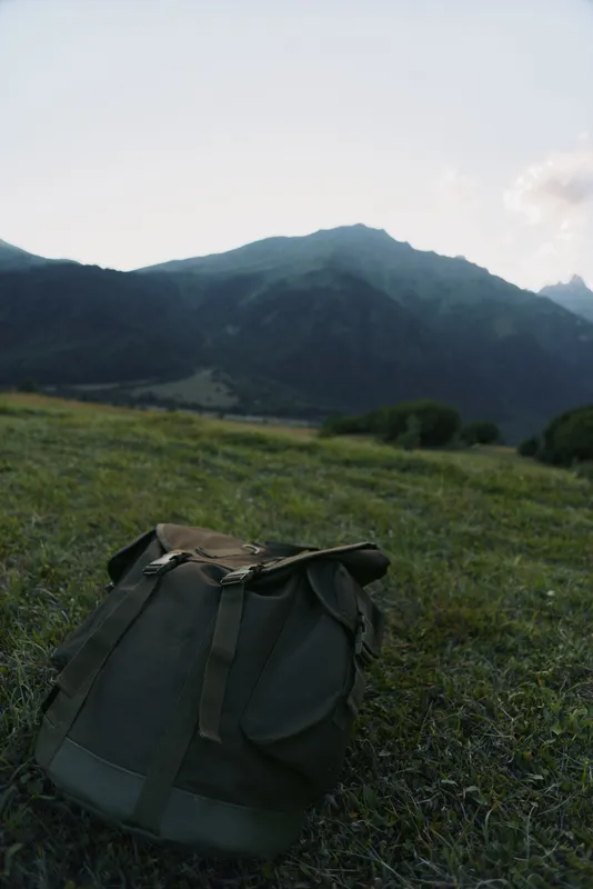 A lone backpack rests on a grassy hillside with distant mountains under a clear sky, inviting exploration and peaceful outdoor solitude.