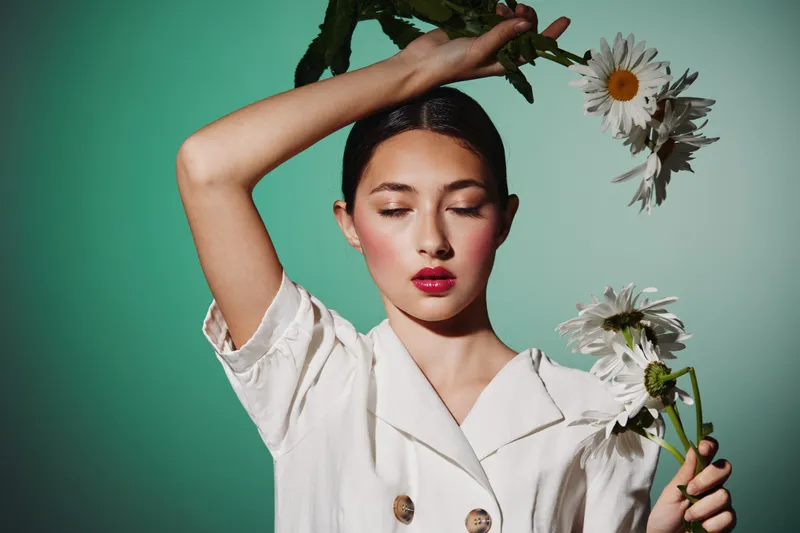 Young woman in white fashion blouse poses with eyes closed and holds white daisies. The creative beauty portrait expresses elegance, softness, and floral inspired fashion style on green background.