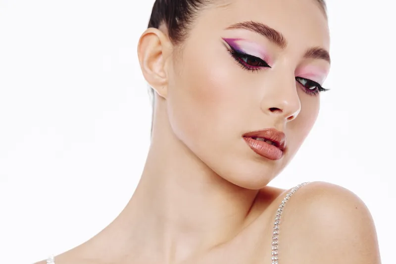 Close-up of a woman with bold colorful makeup featuring vivid pink and purple eyeshadow along with glossy lips. High fashion beauty concept on a white background.