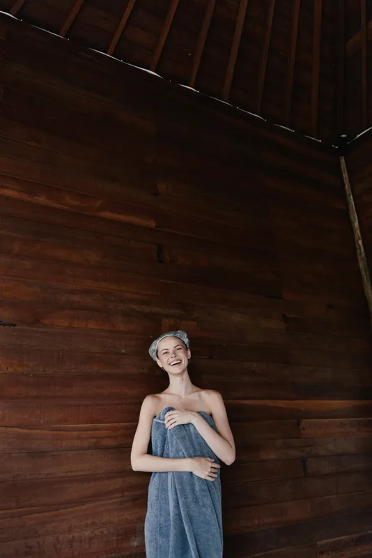 smiling woman wrapped in towel with headband standing against wooden wall in spa environment, relaxed mood and natural lighting, wellness concept